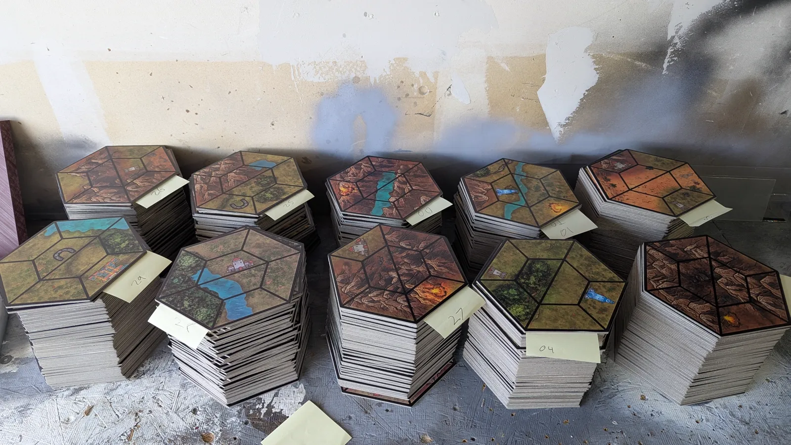 UV-printed game boards, modular hex tiles and tokens by Kulworks: navy and gold board panels printing on the flatbed and coming off finished, hex terrain tiles cut on the clicker press and freed from the die, a full run stacked and bundled on the bench, double-sided wooden game tokens printed in a jig, and tile artwork prepped for print